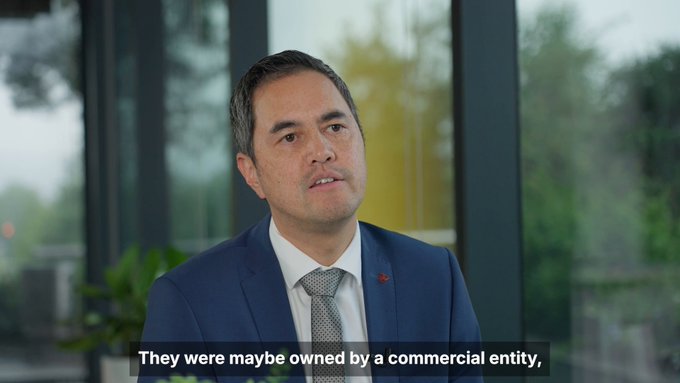 A video interview still of a man in a blue suit and tie speaking, with a caption reading, "They were maybe owned by a commercial entity,"