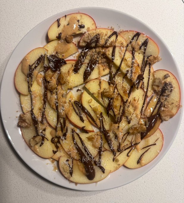 A plate of sliced apples drizzled with peanut butter, chocolate syrup, and what appears to be granola or crushed nuts.