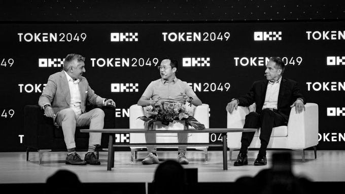 Three panelists engage in a fireside chat discussion on the OKX Main Stage at TOKEN2049, seated in white chairs with a branded backdrop displaying "OKX" and "TOKEN2049" logos. The accompanying post highlights the event's focus on building trust between regulators and industry leaders in shaping the future of digital finance, with the middle panelist appearing to be Star Xu, OKX's Founder and CEO.