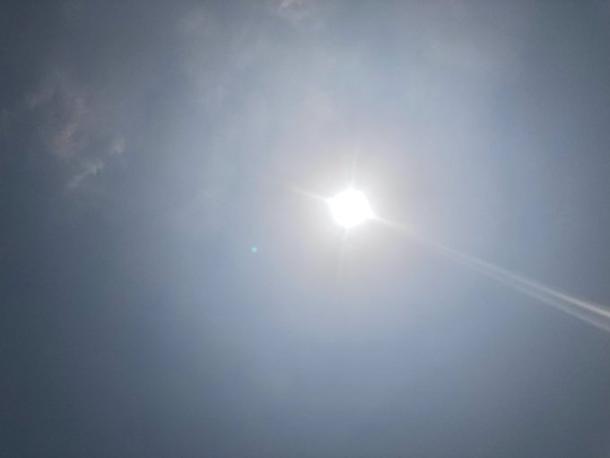 A bright sun shining in a pale blue sky with wispy clouds and a faint contrail visible.