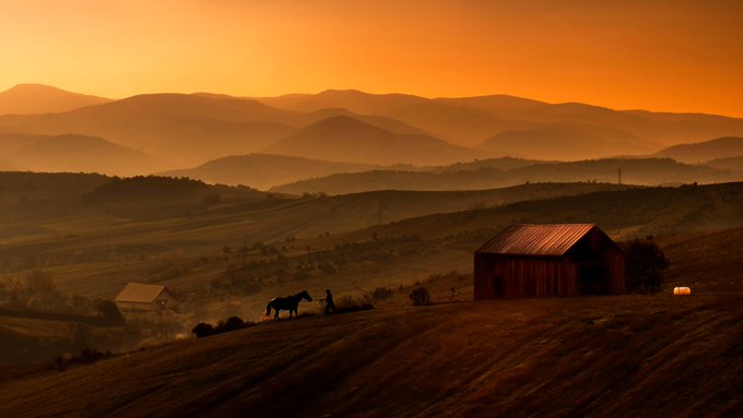 The image features a horse standing next to a red barn, with mountains visible in the background under an orange sky. The scene appears to be set in a rural area or countryside setting, possibly near a farm.