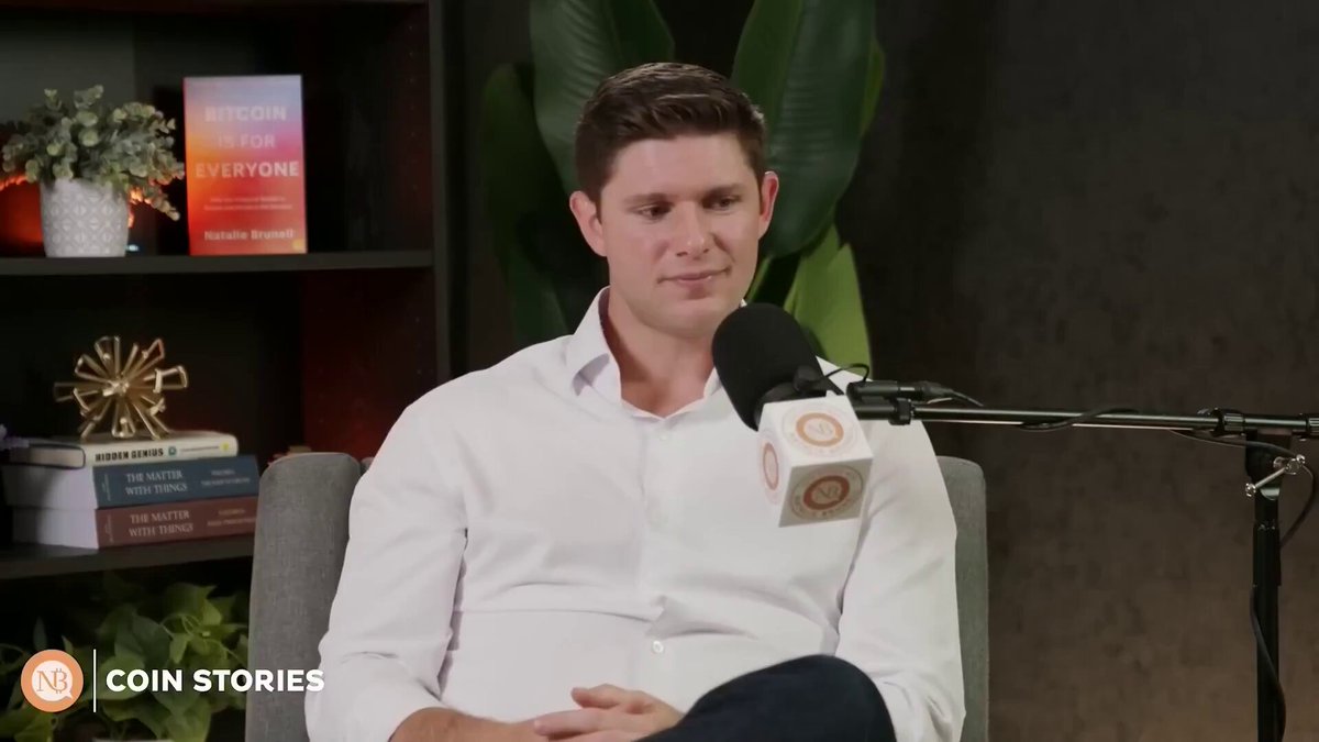 The image shows a man sitting in a chair, wearing a white shirt. He has a microphone in front of him, suggesting that he might be hosting or participating in an interview. The background features a bookshelf filled with various books, indicating that this could be a library or a reading area. There is also a potted plant and a vase on the shelf, adding to the ambiance of the space.