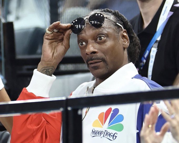 Snoop Dogg wearing a white jacket with an NBC 'Snoop Dogg' logo, lifting his sunglasses to make a surprised wide-eyed expression while sitting in a venue.