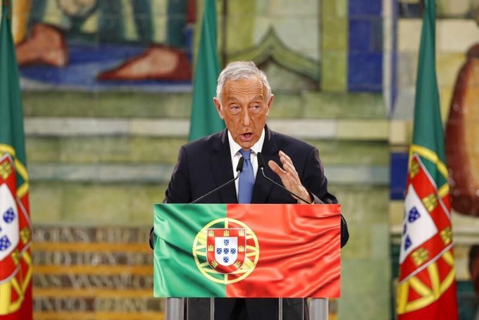 The image features a man giving a speech or lecture, likely at an event related to Portuguese culture, politics, or history. The man is standing behind a podium and making his point while wearing a suit and tie. He is looking towards the camera with a concerned expression. In the background, there are flags from different countries, which suggests that this gathering might be of international significance or could be taking place in a multilingual location. A text overlay on the image provides some context about Portuguese tax regulations for cryptocurrency gains if held for one year and offers advice on how to avoid paying tax on these gains.