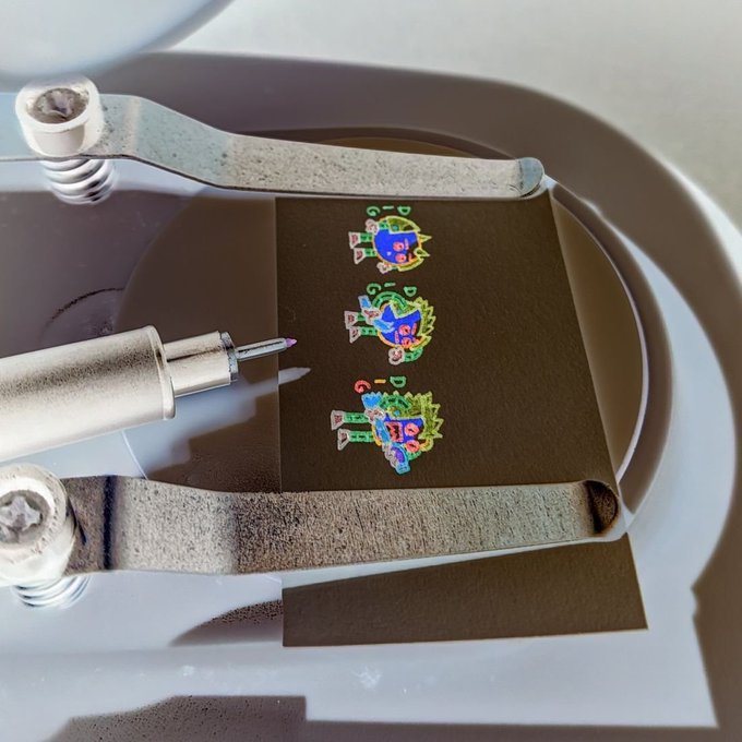 A close-up of a laser engraving device in the process of burning colorful cartoon character designs onto a strip of dark material, with visible smoke and sparks.