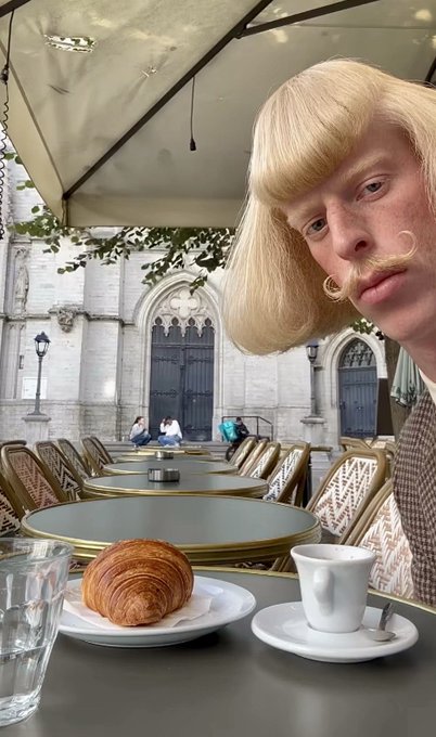 A surreal photo showing a person with a blonde bob haircut and mustache filter taking a selfie at an outdoor café with a croissant and espresso on the table.