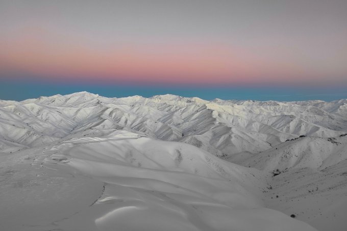 Sweeping snow-covered mountain ridges stretch across the horizon under a pastel pink and blue sky at dusk or dawn.