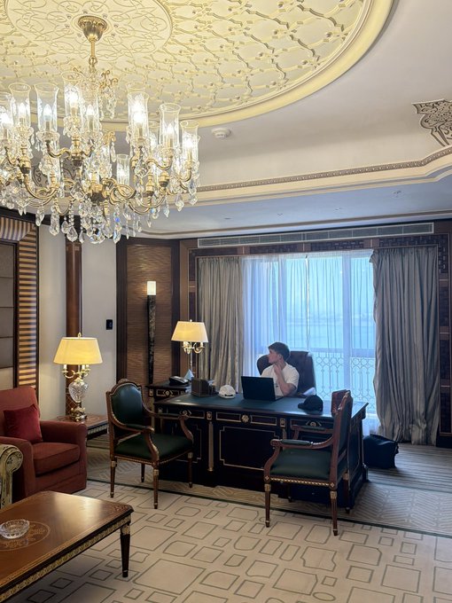 A luxurious hotel room or office with ornate gold ceiling details, a large crystal chandelier, and a person working on a laptop at a dark wood desk.