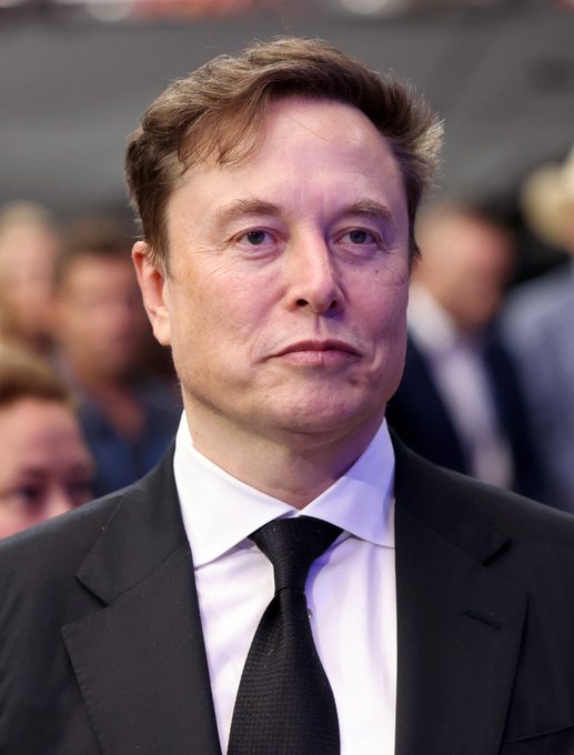 Elon Musk, dressed in a black suit with a white shirt and black tie, stands with a serious expression in a crowded venue. The image aligns with the post's satirical, fawning tone, visually representing the subject of the elaborate praise.
