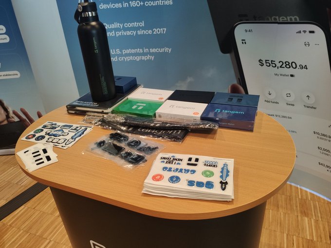 A Tangem booth display at a trade show showing a black water bottle, various branded cards and stickers, and a large mock-up of a mobile app showing a wallet balance of $55,280.94.