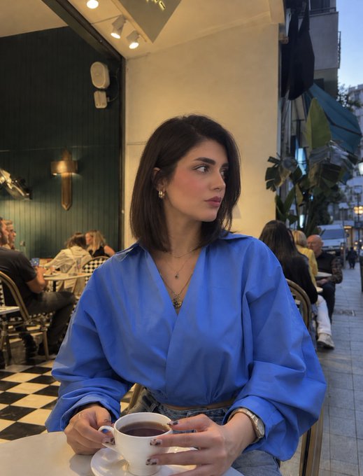 A woman with short brown hair wearing a blue blouse sits at an outdoor cafe table, holding a cup of dark liquid and gazing off to the side.