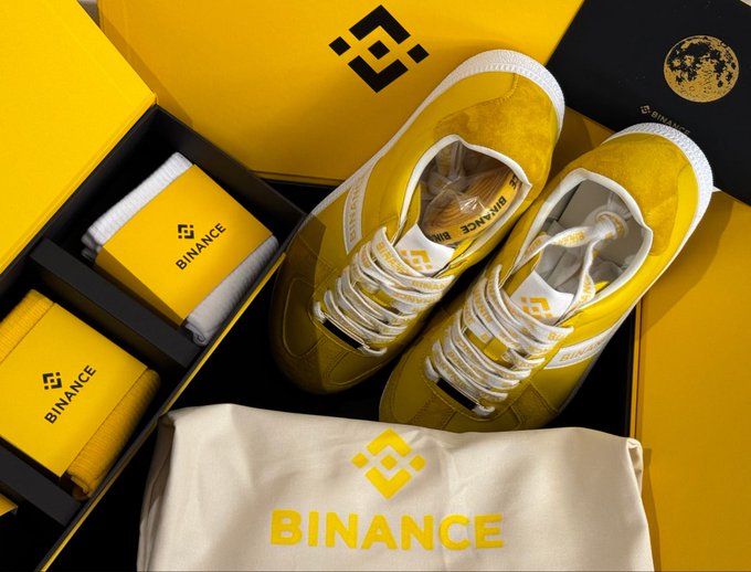A pair of yellow Binance-branded sneakers is displayed alongside yellow socks and a drawstring bag featuring the Binance logo, all nestled in matching yellow packaging boxes. The post invites followers to choose their preferred gift from Binance or MEXC, with the image showcasing one of the promotional items Binance is offering.