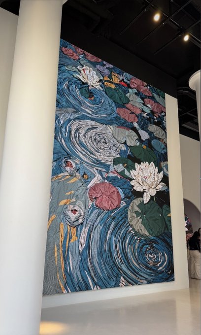 A monumental textile artwork installed in a gallery, depicting swirling blue water, white water lilies, and koi fish, with the Instagram caption "scale matters" highlighting how the massive size of the piece transforms the intimate subject matter into an immersive experience.