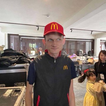 A young man wearing a red McDonald's cap and dark vest with the yellow 'M' logo, standing indoors at what appears to be a McDonald's location, smiling at the camera.