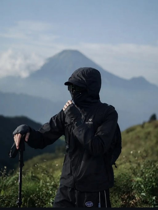 A person dressed in a black hooded Arc'teryx jacket, face mask, and sunglasses, holding a hiking stick in a mountainous landscape.