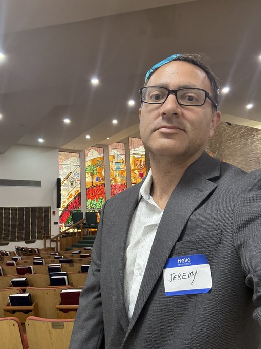 A man wearing a gray suit jacket and a blue yarmulke has a 'Hello My name is Jeremy' name tag, standing in a synagogue with stained glass windows behind him.