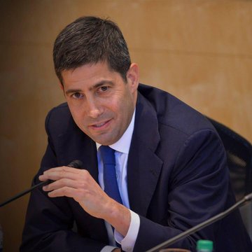 The image features a man dressed in a suit and tie sitting at a desk, with his hand resting on his chin as he looks off into the distance or at someone else. The background of the image shows a wall that has a green bottle mounted on it. There is also text present in the image, which reads "The new Fed Chair Kevin Warsh is pro-Bitcoin. If you’re under 40, Bitcoin is your new Gold." This suggests that the man might be discussing or advocating for Bitcoin as an investment option.