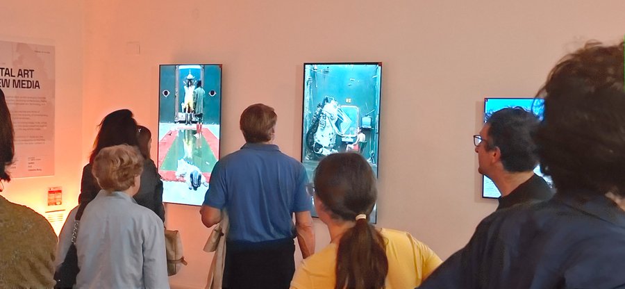 A group of people standing in an art gallery watching video installations displayed on vertical screens, with a wall text partially visible reading 'ITAL ART NEW MEDIA'.