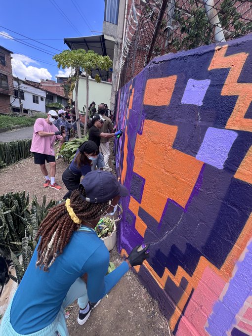 A group of volunteers collaborates on painting a vibrant geometric mural in purple, orange, and pink hues along a community wall, with cacti and lush hillside homes in the background. The scene embodies the post's message of participatory art revitalizing neighborhoods, reflecting the resilience and creativity of the Colombian community behind "Primavera Eterna – The Eternal Spring."