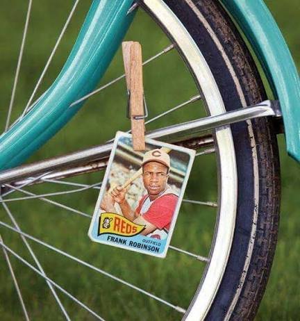 A vintage baseball card of 'Frank Robinson Outfield' of the 'REDS' is clipped to the spokes of a teal bicycle wheel.