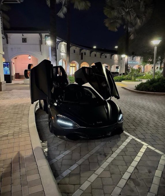 A black McLaren supercar with its dihedral doors open, parked on a paved area at night in front of a building with arched architecture and palm trees.