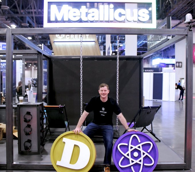 A man sits on a swing inside a booth at a trade show, with a large "Metallicus" sign above. He holds a yellow coin featuring the letter "D" (Dogecoin) and a purple coin featuring an atom symbol (Cosmos).