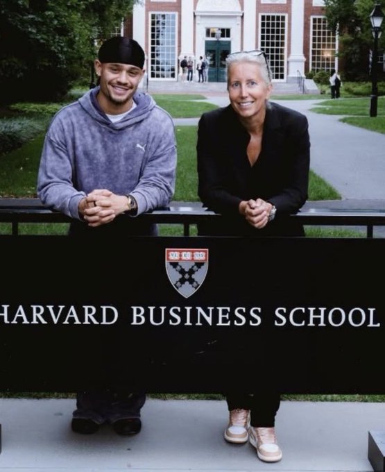 Two people pose smiling behind a black "HARVARD BUSINESS SCHOOL" sign featuring the university's shield emblem, with one being footballer Xavi Simons who has enrolled in the business program to broaden his knowledge beyond football.