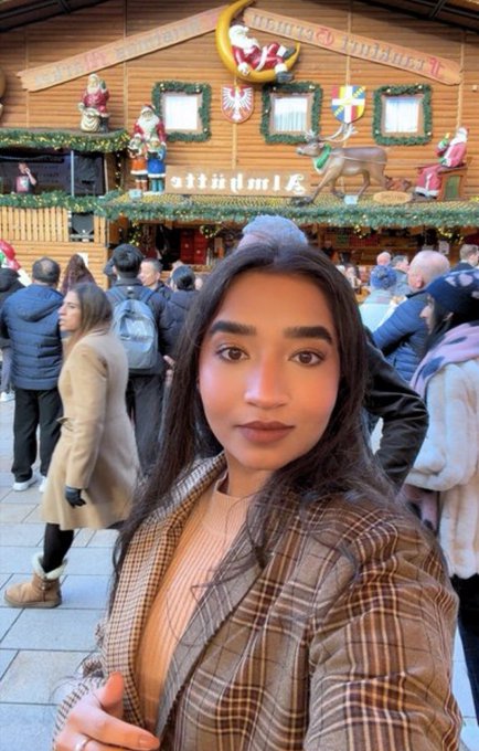 A woman in a checkered coat takes a selfie in front of a wooden market stall decorated with Christmas-themed ornaments and a Santa figure.