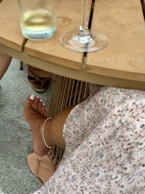 A first-person view looking down at a person's feet in sandals under a table with wine glasses, with the watermark '@WyonaWyld' visible.