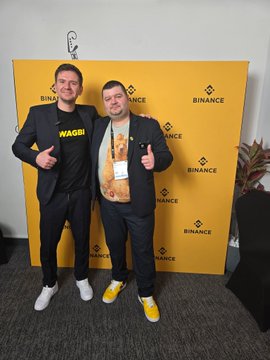 Two men pose in front of a yellow Binance branded backdrop; one wears a black t-shirt with yellow "WAGMI" text.