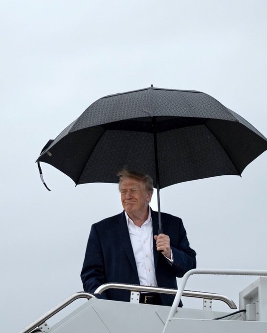 A man in a dark suit and white shirt holds a black umbrella while descending the stairs of an aircraft against a gray sky.
