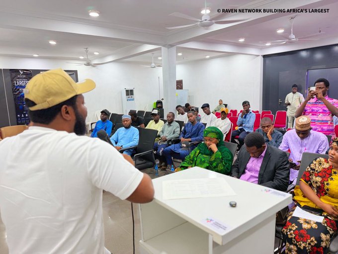 A man in a white shirt and yellow cap speaks into a microphone to a seated audience in a conference room, with text reading '© RAVEN NETWORK BUILDING AFRICA'S LARGEST'.