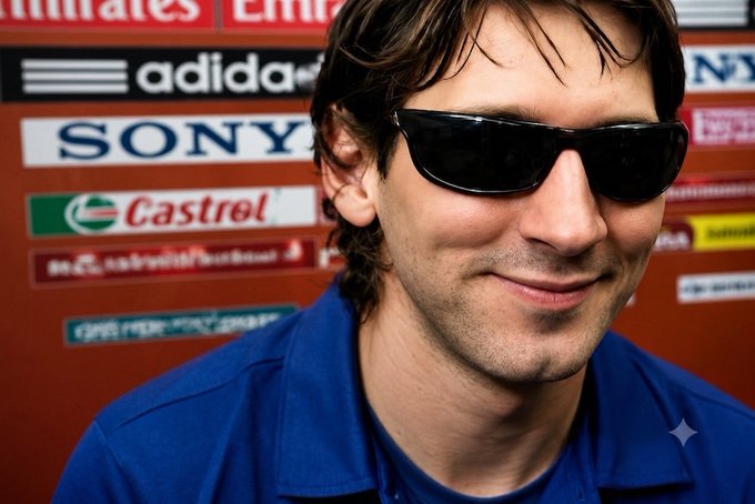 A young man with dark hair wearing black sunglasses and a blue collared shirt smiles at the camera, standing in front of a backdrop featuring sponsor logos including Emirates, Adidas, Sony, and Castrol.
