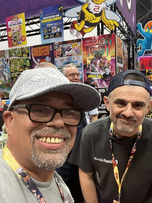 Two men smile for a selfie at a comic convention, with booths for 'VIZ', 'WEBTOON', and 'KODANSHA COMICS' visible in the background.