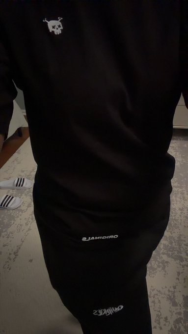 A person wearing a black matching set with a small white bull logo on the chest and 'ORIGINALS' text on the pants, standing on a carpet with white slides visible nearby.