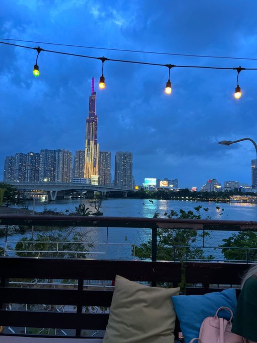 A waterfront view at dusk captures a tall illuminated skyscraper across a river, framed by string lights and cushioned seating on a balcony.