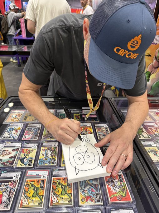 A man wearing a blue 'Chipotle' cap leans over a glass display case filled with trading cards, drawing a cartoon face on a white autograph booklet with a black marker. He wears a lanyard with a credential around his neck.