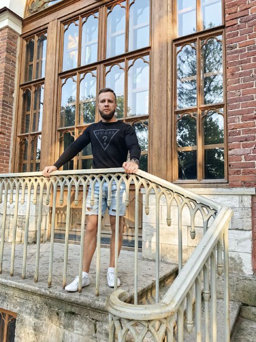 A man wearing a black "GUESS" long-sleeve shirt and denim shorts stands confidently on a Gothic-style stone staircase, resting his hands on an ornate metal railing in front of tall arched wooden windows. The image complements the post's message by visually representing someone who has persevered through a tough week and made it to Friday, standing tall and composed despite the "fight" referenced in the caption.