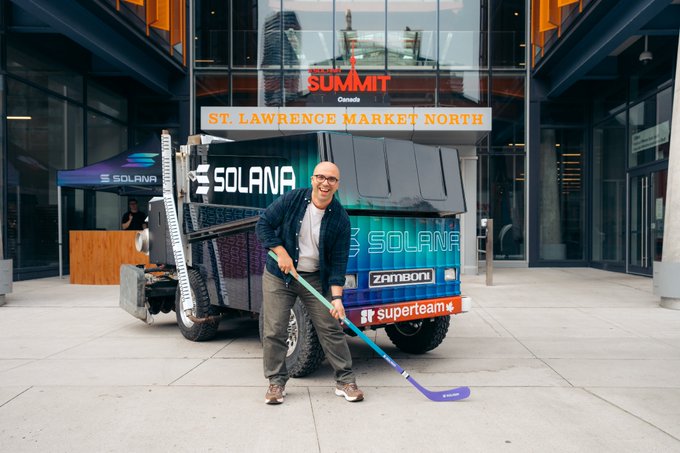 A bald man in a suit poses with a hockey stick next to a teal Zamboni branded with 'SOLANA' and 'superteam' logos outside the St. Lawrence Market North building during what appears to be a Summit Canada event.