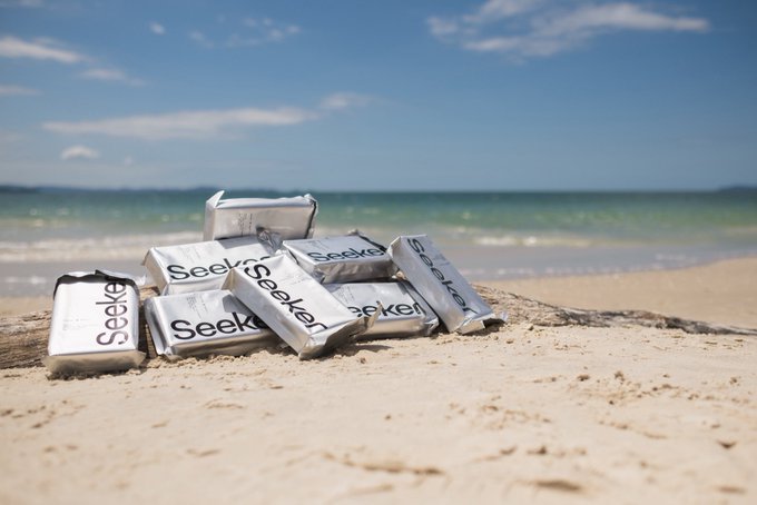 A pile of silver Seeker-branded phone packages rests on a sandy beach with the ocean in the background. The image illustrates the post’s announcement that Seeker Connect is giving Seeker phones to dApp Store builders at Solana Mobile’s Seeker Island.