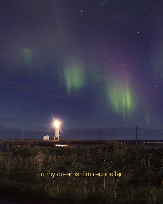 A lighthouse stands illuminated against a night sky lit by green and purple auroras, with the caption "In my dreams, I'm reconciled" overlaid in yellow text at the bottom. The image accompanies a promotion for the photographer's "Keep Me There" series on Transient Labs, with the lighthouse serving as a solitary beacon that echoes themes of longing and reconciliation.