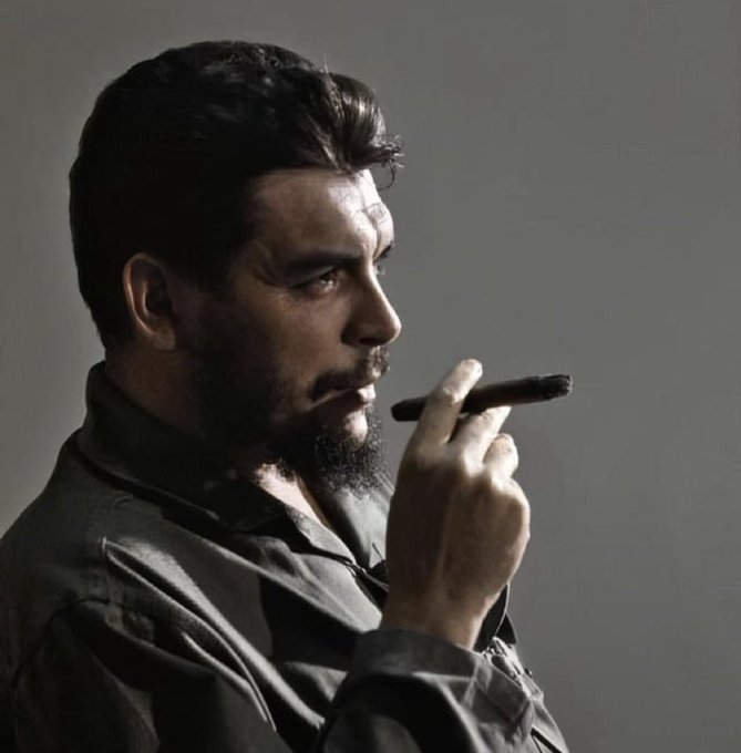 A side-profile portrait of a bearded man with dark hair wearing a dark shirt, smoking a cigar against a plain gray background with dramatic lighting.