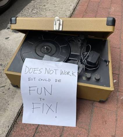 A broken vintage yellow record player sits on a sidewalk with a handwritten sign that reads "DOES NOT WORK BUT COULD BE FUN TO FIX!".
