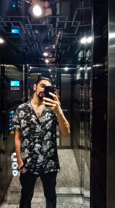 A mirror selfie of a man with a beard wearing a black Hawaiian-style shirt with white patterns, standing in a mirrored elevator.