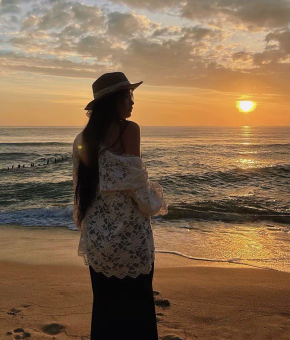 A woman with long dark hair wearing a tan hat and a white lace coverup looks out at a golden sunset over the ocean from a sandy beach.