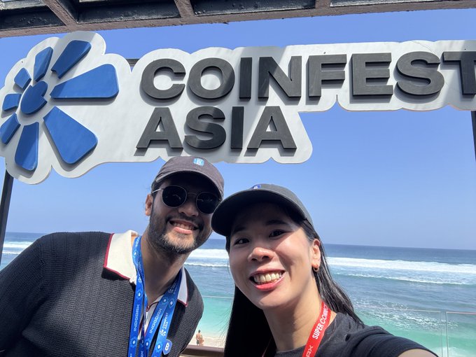 A selfie of a man and a smiling woman at 'COINFEST ASIA', both wearing caps and lanyards, with the ocean and blue sky visible in the background.