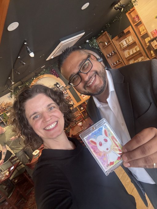 A smiling woman with curly hair and a bearded man wearing glasses pose together indoors, with the man proudly displaying a 1/1 "Reliable Rat" VeeFriends Topps Chrome Card featuring a cartoon rat character. The image captures a memorable Comic Con meetup between the card's owner and its collector, celebrating the community spirit highlighted in the post.