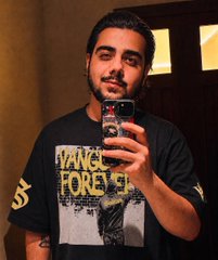 A mirror selfie of a young man with a beard wearing a black 'Tango Forever' t-shirt, holding a smartphone in a warmly lit indoor setting.
