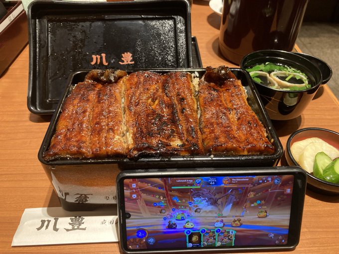 A takeout meal of grilled eel (unagi) rice in a black container with Japanese characters, accompanied by miso soup and pickles, with a smartphone displaying gameplay in the foreground.