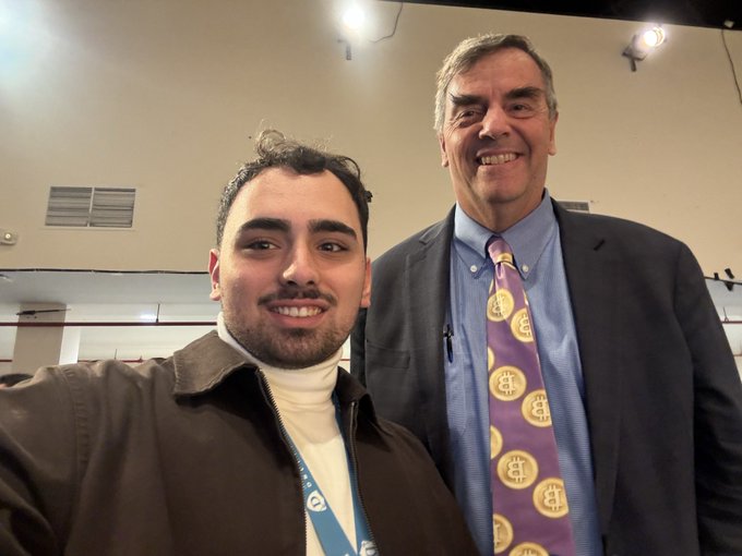 A selfie of two men smiling at the camera; the younger man on the left wears a brown jacket over a white turtleneck with a lanyard, and the older man on the right wears a blue suit with a purple tie featuring a gold circular pattern.